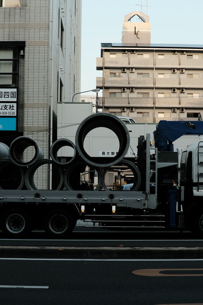 services-03 Truck carrying large pipes on a city street during the day, captured in an urban environment.