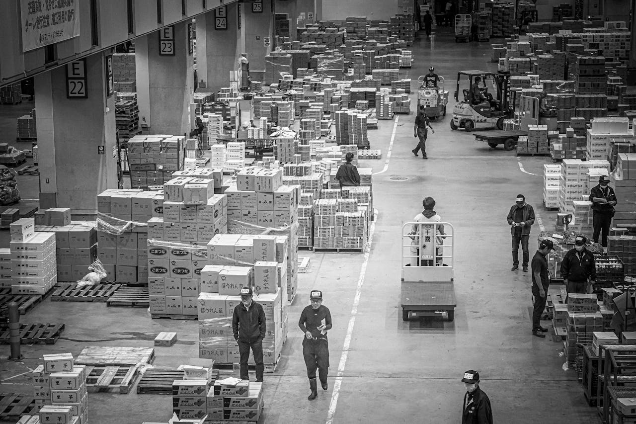services-02 Black and white photograph of a bustling warehouse in Japan, featuring workers organizing and moving packages.