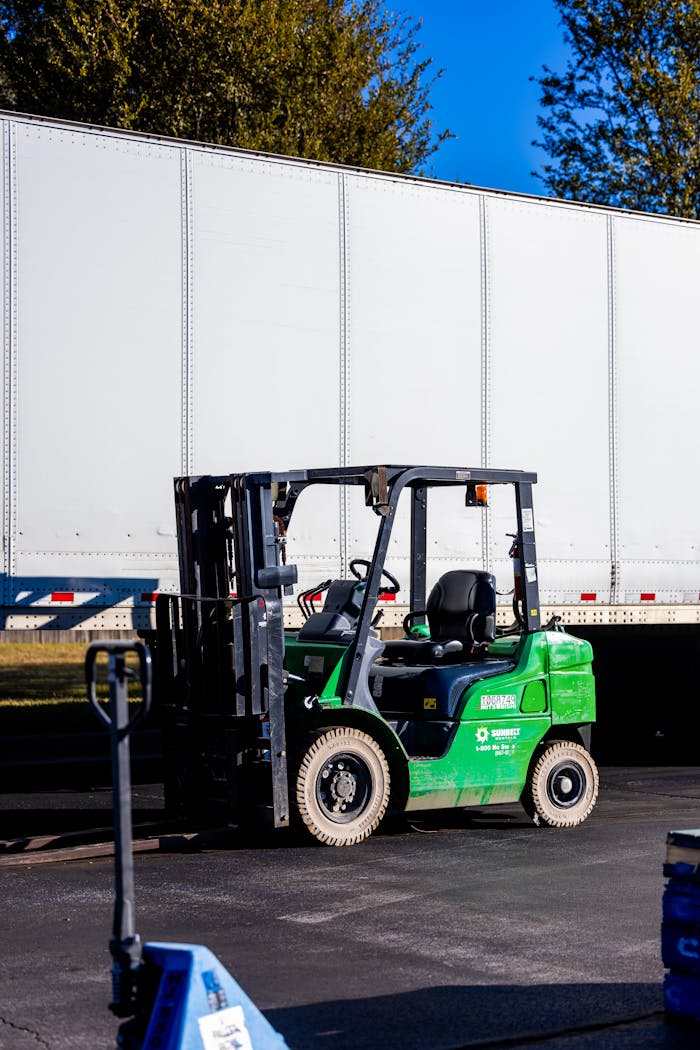 gallery-1 A green forklift parked beside a large white truck outdoors on a clear day.