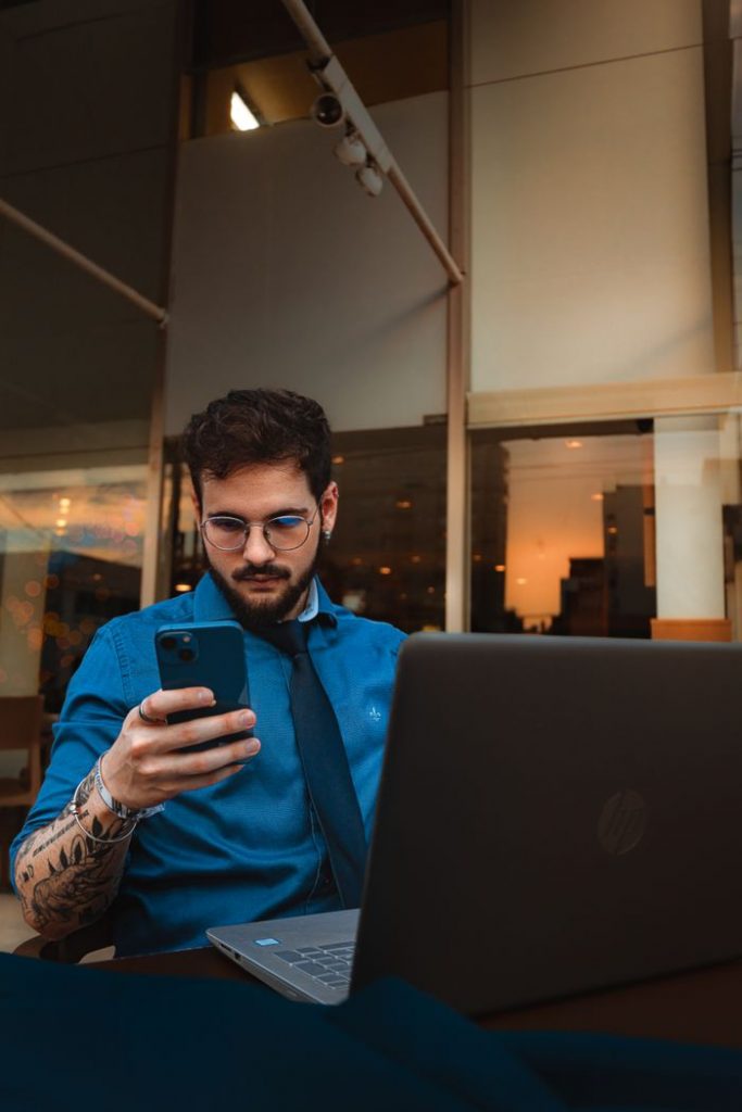 pexels photo 16284923 16284923 A man with a beard and eyeglasses focuses on his smartphone, sitting indoors with a laptop.